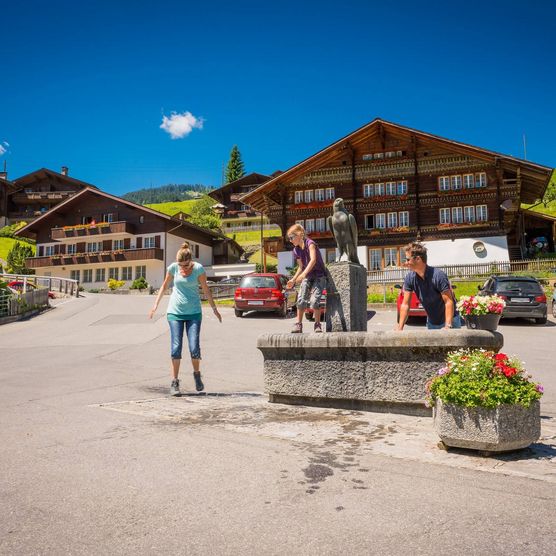 Familie erfrischt sich auf dem Dorfplatz im Brunnen. Rundherum befinden sich traditionelle Chalets und Wohnhäuser.